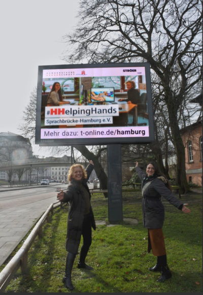 Annja Haehling von Lanzenauer und Susanna Jordanovic von Sprachbrücke-Hamburg e.V. vor einer Stroer-Werbetafel und der #HHelpingHands-Werbung